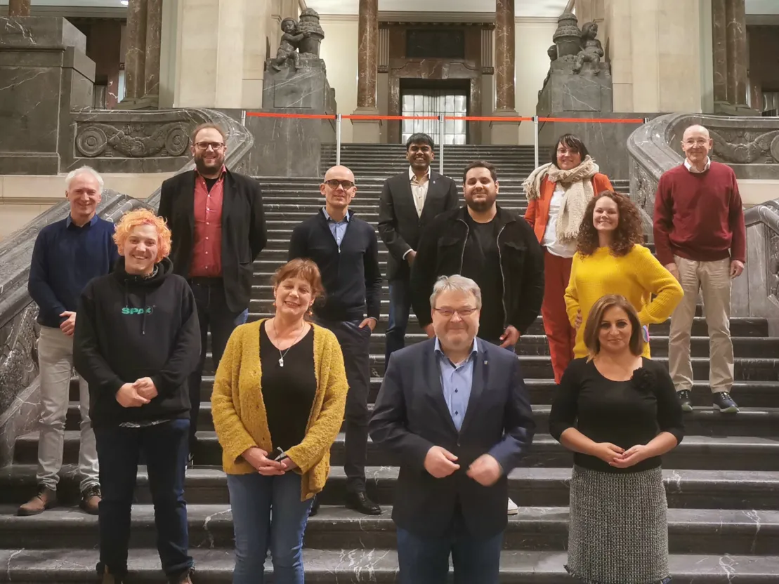 Zw&ouml;lf Menschen, acht M&auml;nner und vier Frauen, stehen in einigem Abstand auf einer gro&szlig;en Freitreppe. Sie posieren f&uuml;r ein Gruppenbild und schauen in die Kamera.